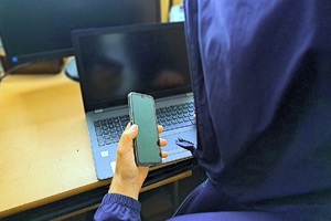 Mężczyzna w kapturze siedzi przed komputerem trzymając telefon w ręku