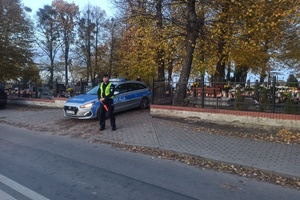 Policjant stoi przy radiowozie, kontroluje ruch pojazdów w rejonie cmentarza